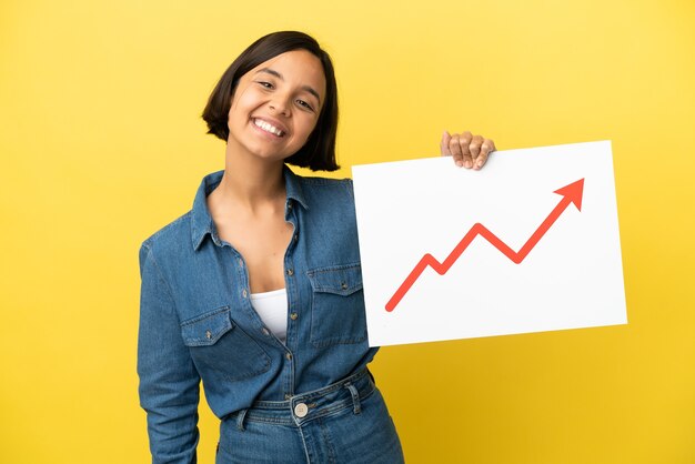 Young mixed race woman isolated on yellow background holding a sign with a growing statistics arrow symbol with happy expression