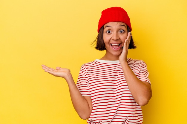 Young mixed race woman isolated on yellow background holds copy space on a palm, keep hand over cheek. Amazed and delighted.