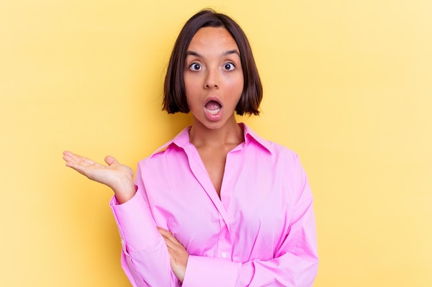 Young mixed race woman isolated on yellow wall impressed holding copy space on palm.