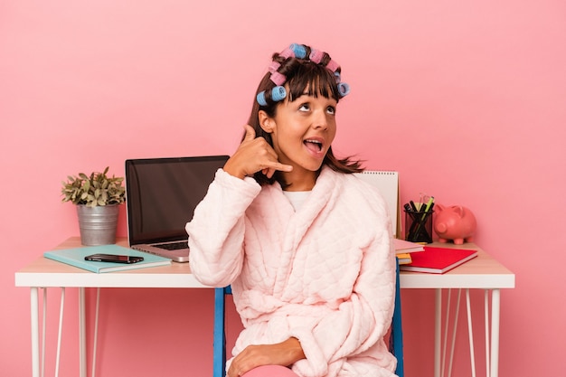 Young mixed race woman in pijama and with curlers isolated on pink background  showing a mobile phone call gesture with fingers.