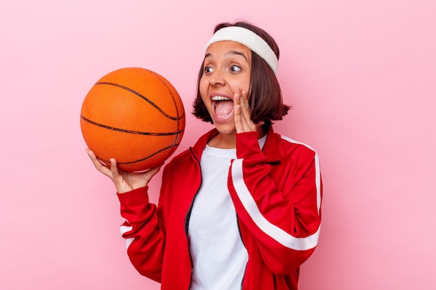 Young mixed race woman playing basketball isolated on pink wall shouting and holding palm near opened mouth.