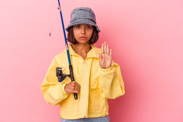 Giovane donna di razza mista che pratica la pesca isolata su sfondo rosa in piedi con la mano tesa che mostra il segnale di stop, impedendoti.