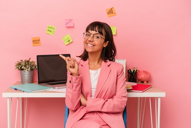 ピンクの背景に分離されたコーヒーを持って部屋で試験を準備している若い混血の女性は笑顔で脇を指して空白のスペースで何かを示しています