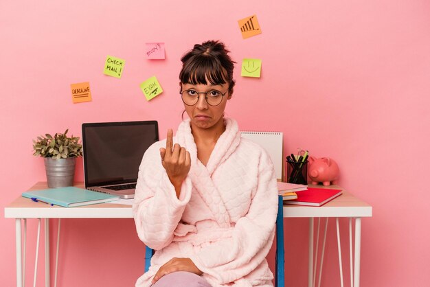 ピンクの背景に分離されたパジャマを着て部屋で試験を準備している若い混血の女性は、招待が近づくようにあなたに指を指しています