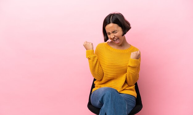 Young mixed race woman sitting on a chair isolated on pink background celebrating a victory
