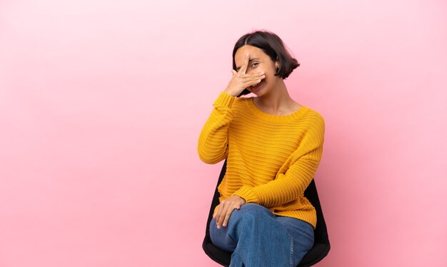 Young mixed race woman sitting on a chair isolated on pink background covering eyes by hands and smiling