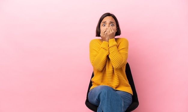 Young mixed race woman sitting on a chair isolated on pink background covering mouth with hands