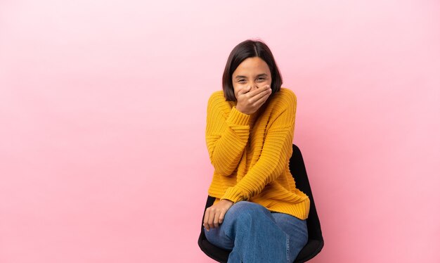 Giovane donna di razza mista seduta su una sedia isolata su sfondo rosa felice e sorridente che copre la bocca con le mani