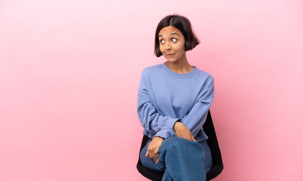 Young mixed race woman sitting on a chair isolated on pink background having doubts while looking up