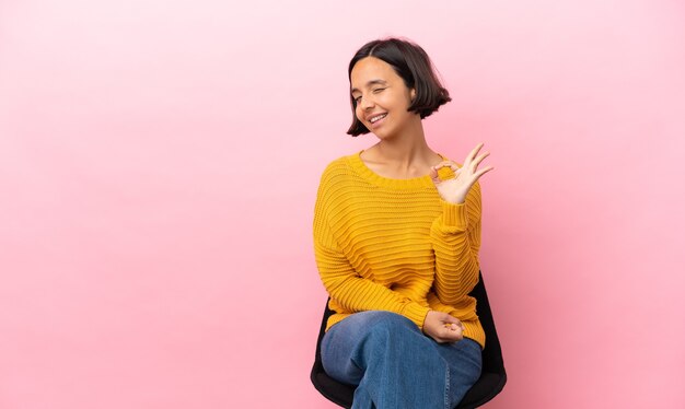 Young mixed race woman sitting on a chair isolated on pink background showing ok sign with fingers