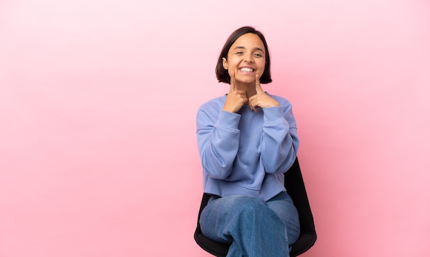 Young mixed race woman sitting on a chair isolated on pink background smiling with a happy and pleasant expression