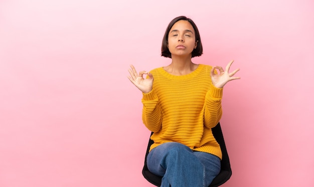 Young mixed race woman sitting on a chair isolated on pink background in zen pose