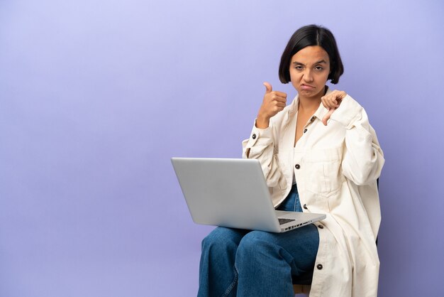 Young mixed race woman sitting on a chair with laptop isolated on purple background making good-bad sign. Undecided between yes or not