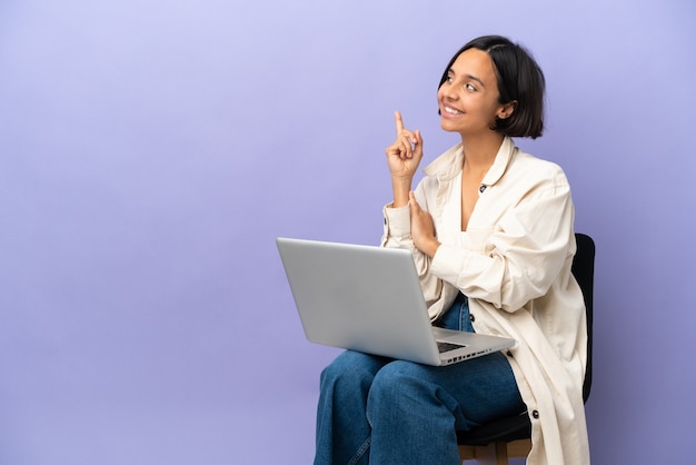 Young mixed race woman sitting on a chair with laptop isolated on purple background pointing up a great idea