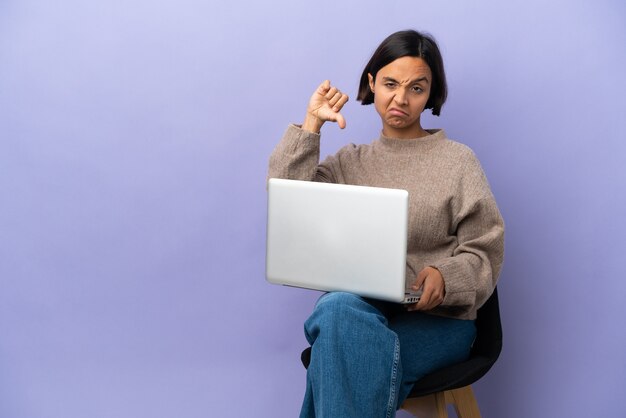 Молодая женщина смешанной расы, сидящая на стуле с ноутбуком, изолирована на фиолетовом фоне, показывая большой палец вниз с отрицательным выражением лица