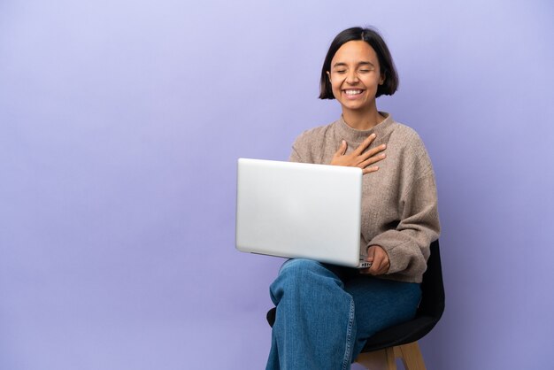 Young mixed race woman sitting on a chair with laptop isolated on purple background smiling a lot