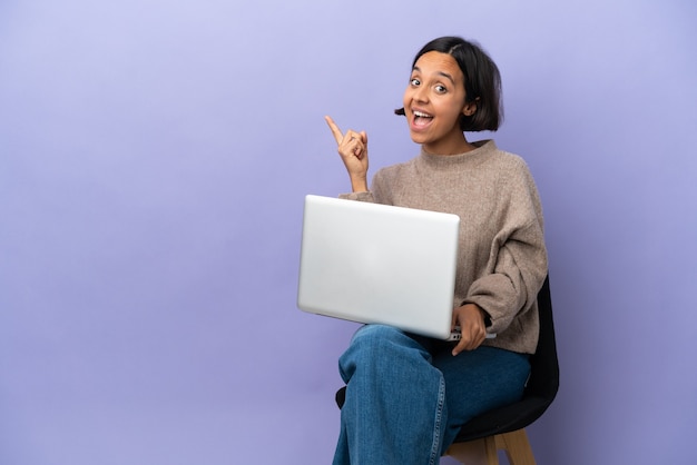 Young mixed race woman sitting on a chair with laptop isolated on purple background surprised and pointing side