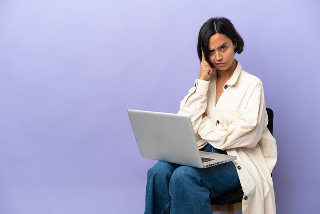 Молодая женщина смешанной расы сидит на стуле с ноутбуком, изолированным на фиолетовом фоне, думая об идее