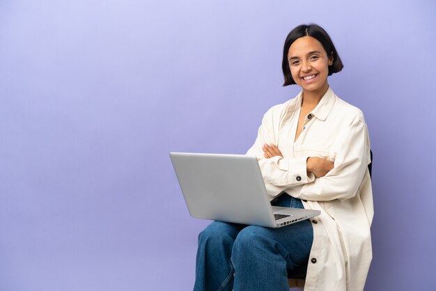 Young mixed race woman sitting on a chair with laptop isolated on purple background with arms crossed and looking forward