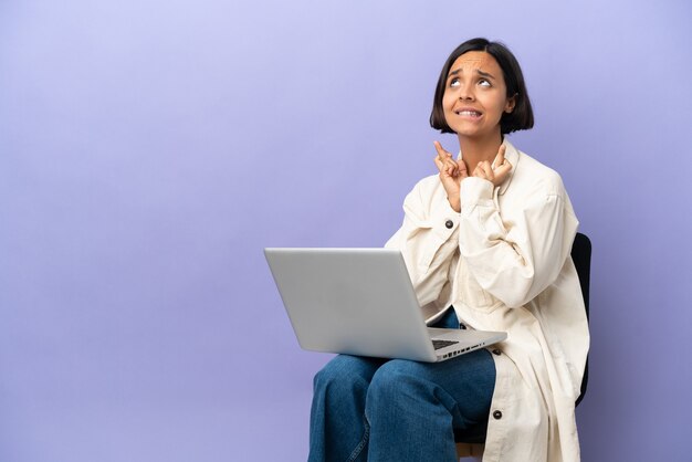 Young mixed race woman sitting on a chair with laptop isolated on purple background with fingers crossing and wishing the best