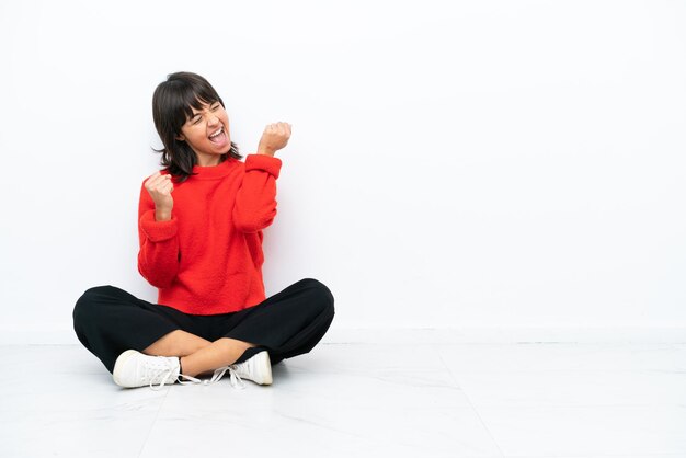 Young mixed race woman sitting on the floor isolated on white background celebrating a victory