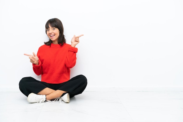 Молодая женщина смешанной расы, сидящая на полу, изолированная на белом фоне, указывая пальцем на боковые стороны и счастливая