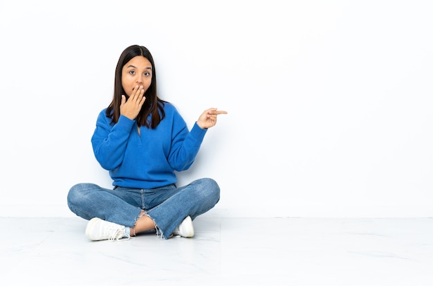 Young mixed race woman sitting on the floor isolated on white background with surprise expression while pointing side