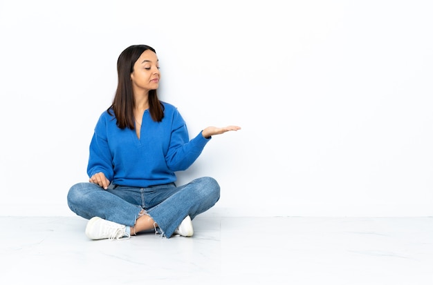 Young mixed race woman sitting on the floor isolated on white  holding copyspace with doubts