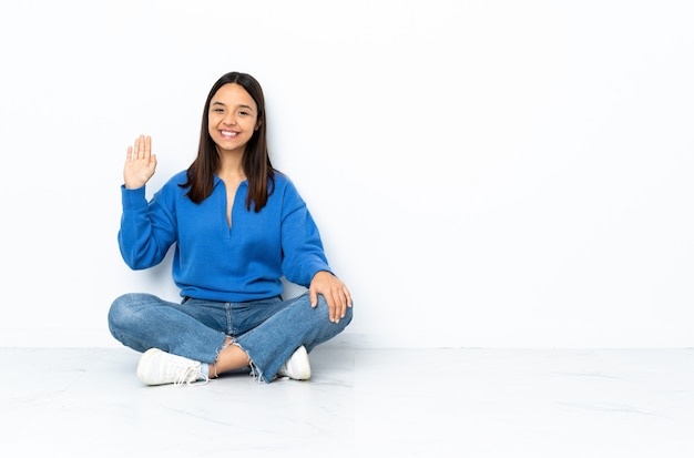 Young mixed race woman sitting on the floor isolated on white saluting with hand with happy expression