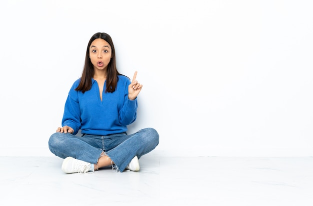 Young mixed race woman sitting on the floor isolated on white space intending to realizes the solution while lifting a finger up