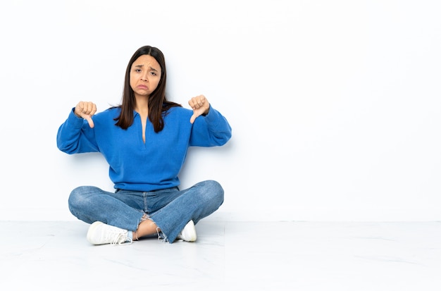 Young mixed race woman sitting on the floor isolated on white wall showing thumb down with two hands