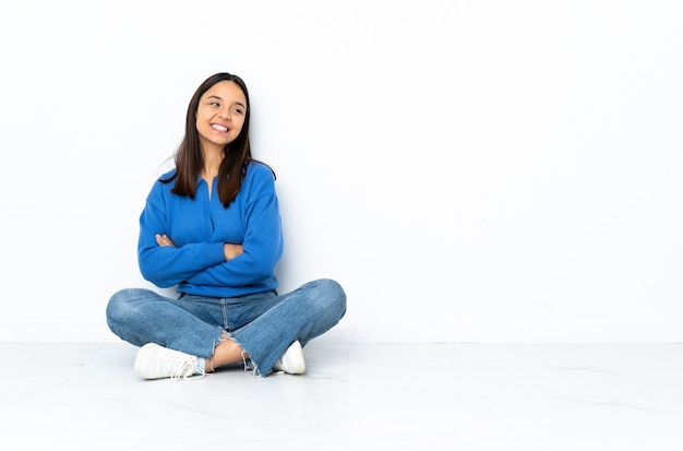 Young mixed race woman sitting on the floor isolated on white wall with arms crossed and happy