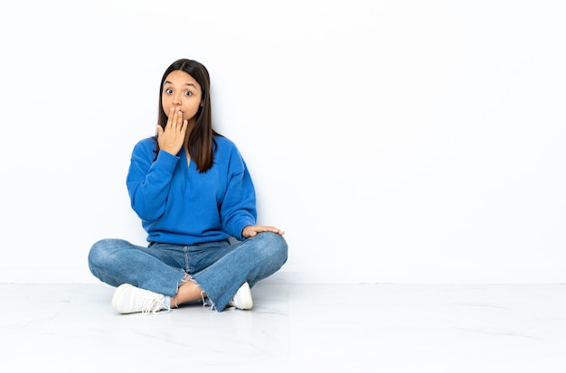 Young mixed race woman sitting on the floor on white wall covering mouth with hand