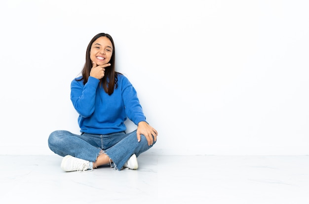 Young mixed race woman sitting on the floor on white wall happy and smiling