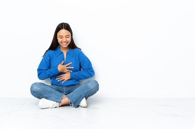 Young mixed race woman sitting on the floor on white wall smiling a lot