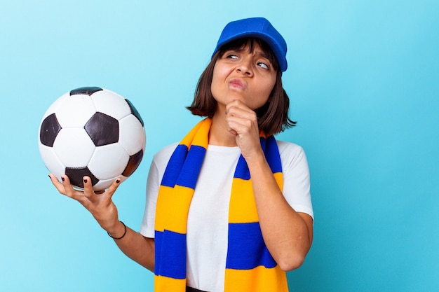 Фото Молодая женщина смешанной расы смотрит футбол, изолированные на синем фоне, глядя в сторону с сомнительным и скептическим выражением лица.