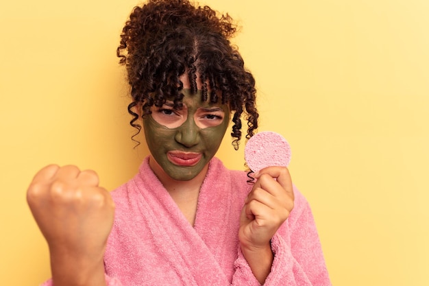 Young mixed race woman wearing a bathrobe holding a makeup remover sponge isolated on yellow background showing fist to camera aggressive facial expression