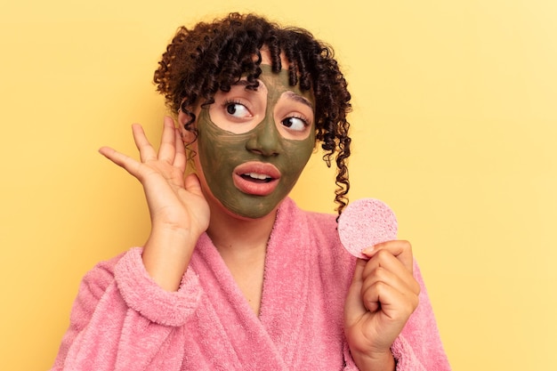 Young mixed race woman wearing a bathrobe holding a makeup remover sponge isolated on yellow background trying to listening a gossip