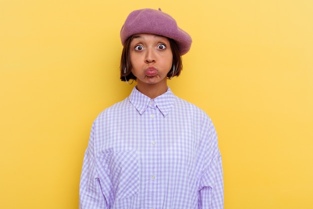 Young mixed race woman wearing a beret isolated on yellow background blows cheeks, has tired expression. Facial expression concept.