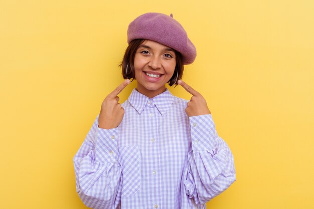 Young mixed race woman wearing a beret isolated on yellow background smiles, pointing fingers at mouth.