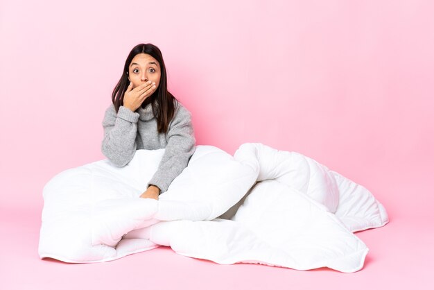 Young mixed race woman wearing pijama sitting on the floor covering mouth with hand