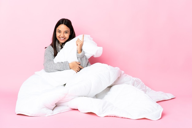 Young mixed race woman wearing pijama sitting on the floor happy and pointing up