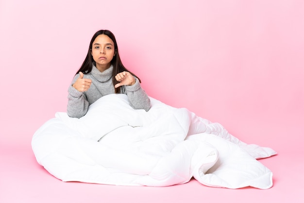 Young mixed race woman wearing pijama sitting on the floor making good-bad sign. Undecided between yes or not