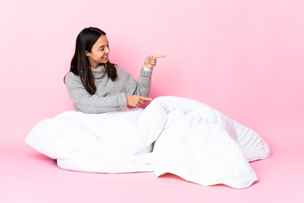 Young mixed race woman wearing pijama sitting on the floor pointing finger to the side and presenting a product