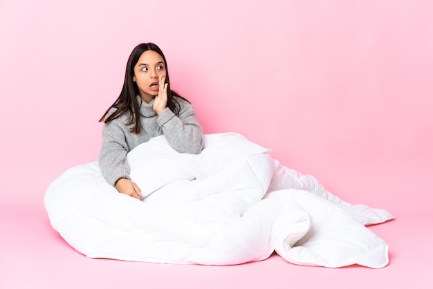 Young mixed race woman wearing pijama sitting on the floor whispering something with surprise gesture while looking to the side