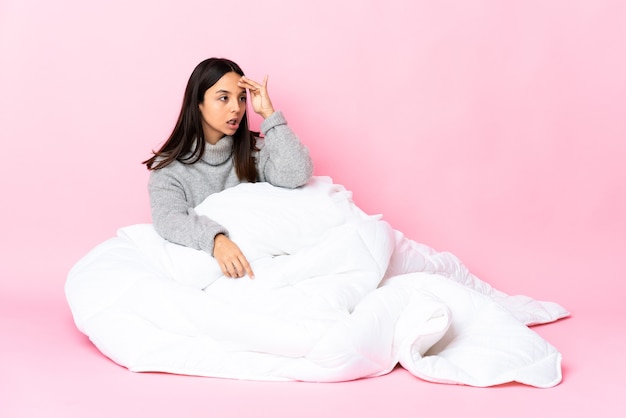 Young mixed race woman wearing pijama sitting on the floor with surprise expression while looking side