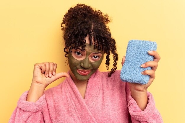 Young mixed race woman wearing a pink bathrobe holding a shower sponge isolated on yellow background feels proud and self confident example to follow