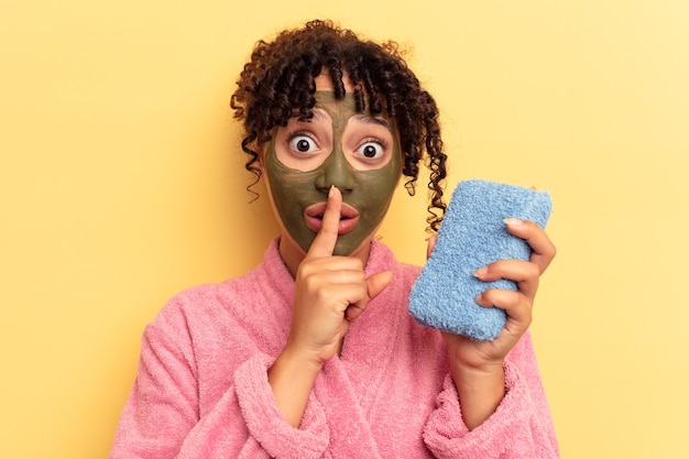 Young mixed race woman wearing a pink bathrobe holding a shower sponge isolated on yellow background keeping a secret or asking for silence.