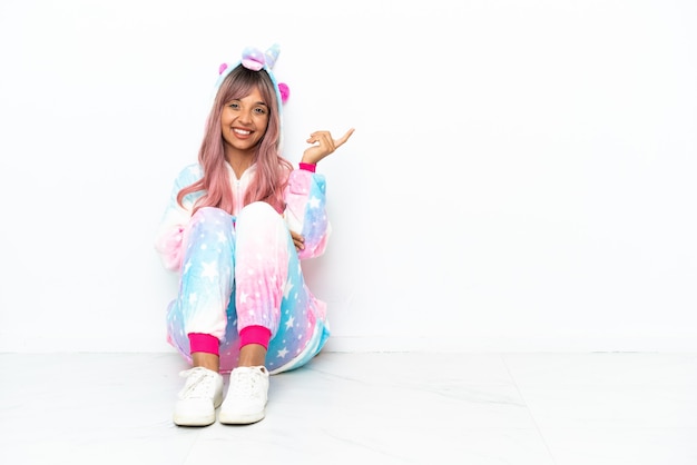 Young mixed race woman wearing a unicorn pajama sitting on the floor isolated on white background extending hands to the side for inviting to come