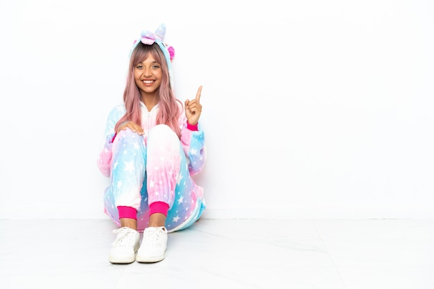 Young mixed race woman wearing a unicorn pajama sitting on the floor isolated on white background pointing with the index finger a great idea